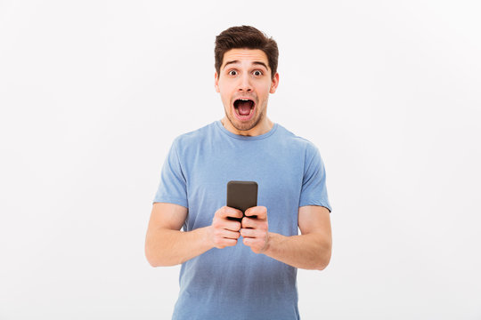 Photo of optimistic guy 30s in casual t-shirt yelling in joy while using mobile phone, isolated over white background