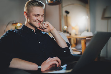 Side view of happy smiling handsome businessman working at night from home, smiling male freelancer working online at vintage loft interior using modern laptop computer