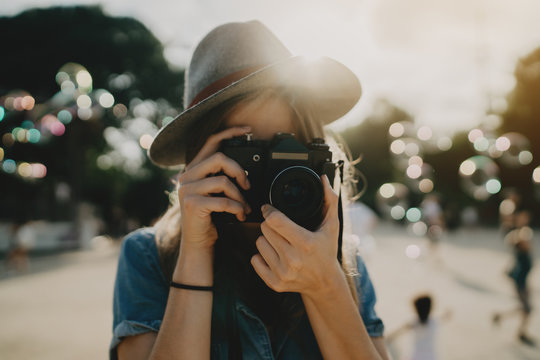 Young hipster girl taking photo on a vintage film camera while walking in the sunny park, stylish woman photographer wearing hat enjoying holidays