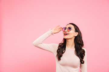 Portrait of elegant woman with red lips wearing round sunglasses smiling and looking upward on copy space, isolated over pink background