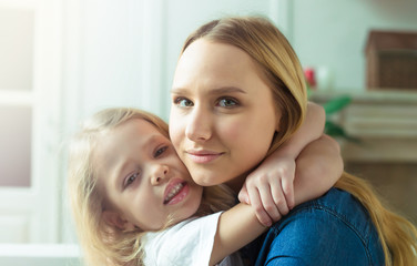 Beautiful smiling blond mother and daughter embrace and have fun on the couch at home and look at the camera. Concept of mother and daughter.