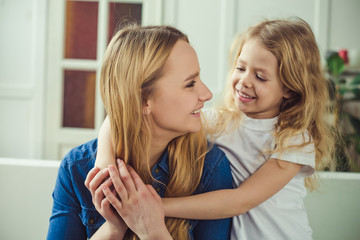 Smiling and happy mom and daughter are hugging each other at home on the couch. Mom and daughter  have fun. 