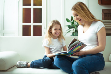 Beautiful smiling blond mother and daughter are sitting on the couch in an embrace. Mom is reading a story to her daughter or a fairy tale from a book.