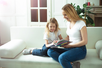 Beautiful smiling blond mother and daughter are sitting on the couch in an embrace. Mom is reading a story to her daughter or a fairy tale from a book.