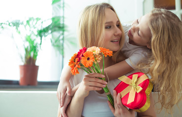 Happy family. Little beautiful smiling girl hugging her mother closing her eyes and gives a gift in a box and a bouquet of flowers. Mothers Day. March 8. Women's Day.
