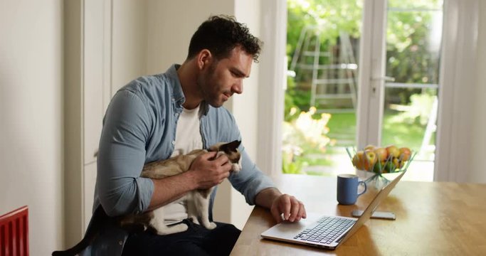 4K Smiling casual man working on laptop at home with his cat sitting on his lap. Slow motion.