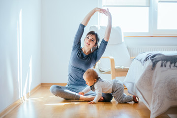 Cute european mother doing yoga or pilates while her infant baby boy playing near at cozy simple home interior. Motherhood, healthy lifestyle and people concept.