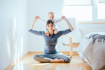 Cute european sporty mother playing with cute infant baby boy. Little son sitting on his mother's shoulders at cozy simple home interior. Motherhood, healthy lifestyle and people concept.
