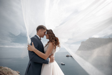 A beautiful bride and groom on their wedding day walk along a rocky shore near the sea.Young Couple On Beach, Happy Smiling Man And Woman Walking Seaside Sea Ocean Holiday Travel. Happy Wedding Day.