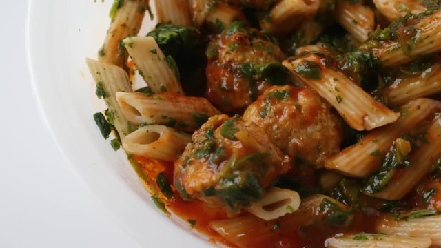 Close slow motion video of a cooked meatball, pasta and spinach in a marinara sauce TV dinner in a plastic tray being stirred with a fork on a white counter top.