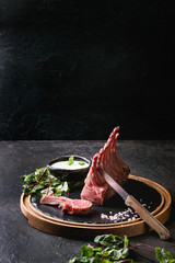 Grilled sliced rack of lamb with yogurt mint sauce served with green salad young beetroot leaves, knife and pink salt on round wooden slate board over dark black table background. Copy space © Natasha Breen