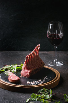 Grilled sliced rack of lamb with yogurt mint sauce served with green salad young beetroot leaves, glass of red wine, pink salt on round wooden slate board over dark black table background. Copy space