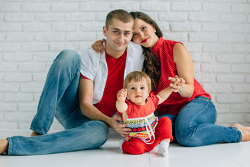 Young happy family sitting on the white floor and having fun. Parents with baby little girl in colorful dress. Man and woman in jeans. Brick wall. Mother father daughter.