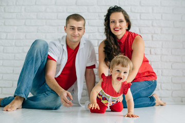Young happy family sitting on the white floor and having fun. Parents with baby little girl in colorful dress. Man and woman in jeans. Brick wall. Mother father daughter.