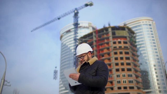 Adult engineer or architect uses a tablet in operation. Writes a message or checks a drawing. Against background is building. Builders are building a modern residential building of glass and concrete