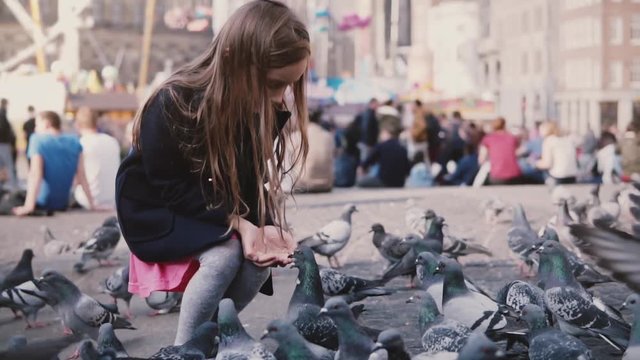 Cute European girl feeding birds from hands. Slow motion. Big flock of pigeons catch seed. Happy little female kid.