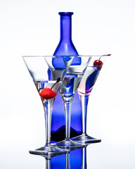 Three martini glasses filled with a cocktail. There is a red cherry in the first and last glass. It is displayed in front of a blue bottle against a white background