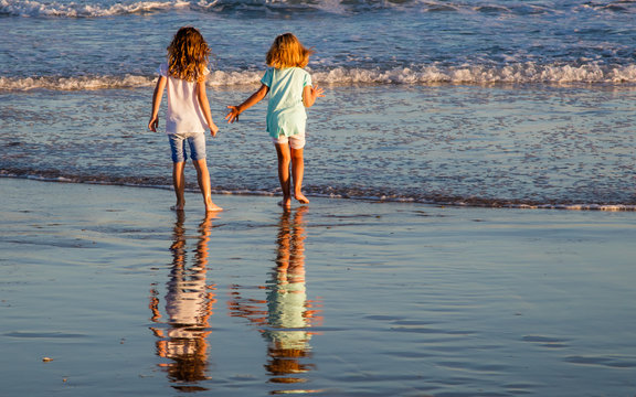 Two little girls playing in the shallow water at the seaside in the late afternoon. Their reflections are visible on the wet sand