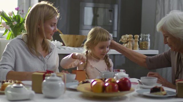 Cute little girl sharing cookies with mother and grandmother and kissing them while having family dinner