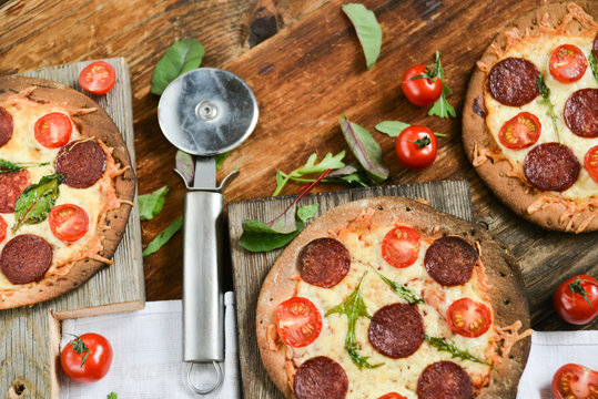 Mini Pizza With Cherry Tomatoes And Salami Sausages On A Wooden Background Natural Rustic, A Pizza Cutter And Ingridienty.