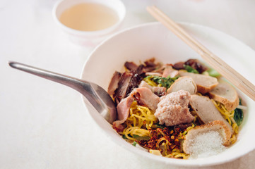 Thai egg noodle in white bowl with sliced red barbecue pork, pork ball, fish sausages close up on white background