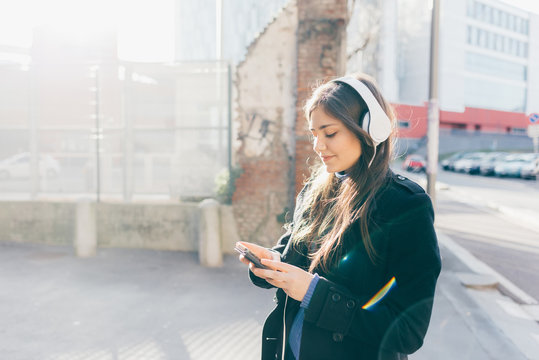 Waist up of young woman posing in the city listening music woth head phones using smart phone hand hold - technology, music, social networking concept