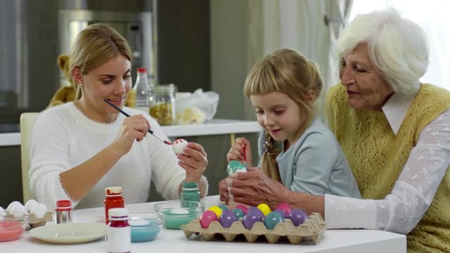Senior lady sitting with daughter and granddaughter at kitchen table and painting eggs for Easter holiday dinner