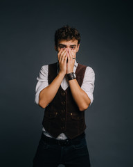 A young, handsome guy posing on a gray studio background. The man is stylish and elegantly dressed. He covers his face with his hands so as not to look at 