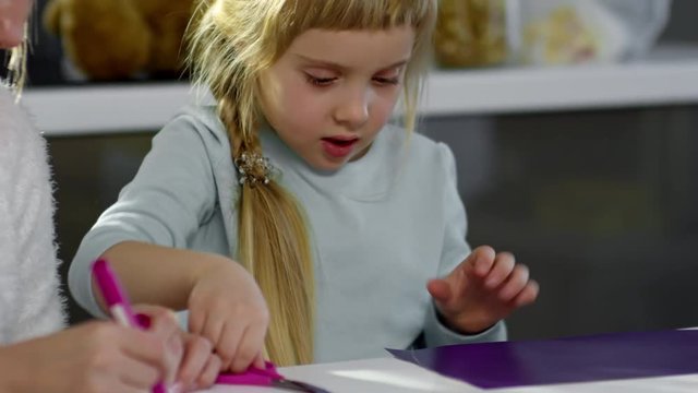 Adorable little girl cutting colorful paper with scissors and talking with mother while she drawing in color book