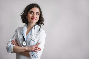 Cute smiling woman with dark curly hair in white medical robe with blue finish stands against a gray wall and looks at the camera. On the neck of a stethoscope.
