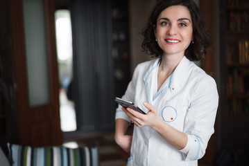 Fototapeta na wymiar Young pretty brunette woman doctor in a white coat holding a phone in hand and smiling. Look at the camera.