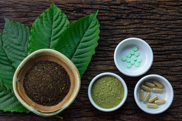 Mitragyna speciosa (kratom) leaves with medicine products in powder, capsules and tablet in white ceramic bowl with wooden texture on background