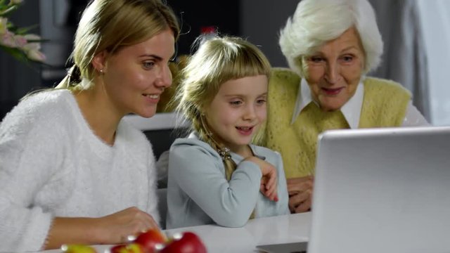 Cute little girl laughing and grimacing at front camera of laptop while sitting with grandmother and mother at table and video calling with family