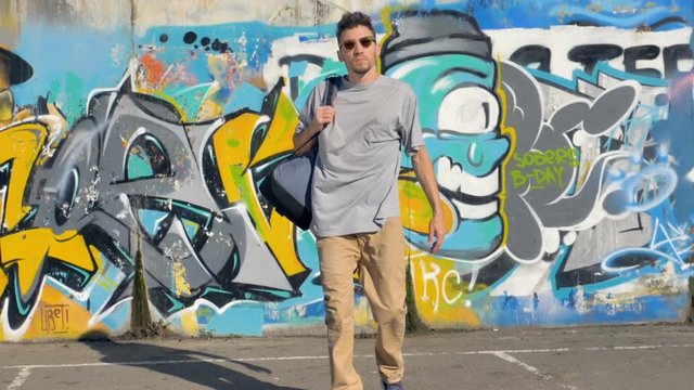 Graffiti artist with a bag on his shoulder is going away from the  wall covered with graffiti pictures.
