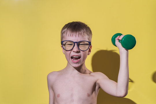 Small boy in glasses holding dumbbell in hand. Child's face feeling very heavy. Funny sport concept.