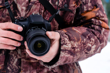 The photographer in a warm camouflage jacket holds the camera with his hands in the winter