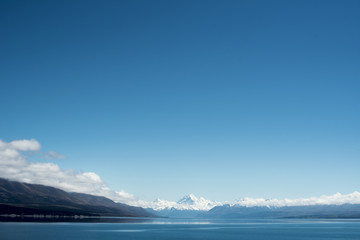 Fototapeta na wymiar Paisaje de picos de montañas nevados con cielo azul y nubes frente un lago y valle con árbol en el Monte Cook, Nueva Zelanda.