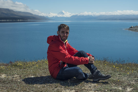 Retrato de hombre joven frente a paisaje de picos de montañas nevados con cielo azul y nubes frente un lago y valle con árbol en el Monte Cook, Nueva Zelanda.