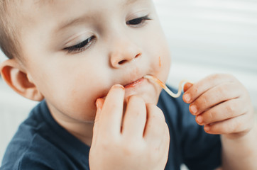 A beautiful cheerful child eats pasta with his hands, spaghetti all dirty
