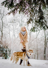 A pleasing blonde in a fur coat stands in the park with her dog