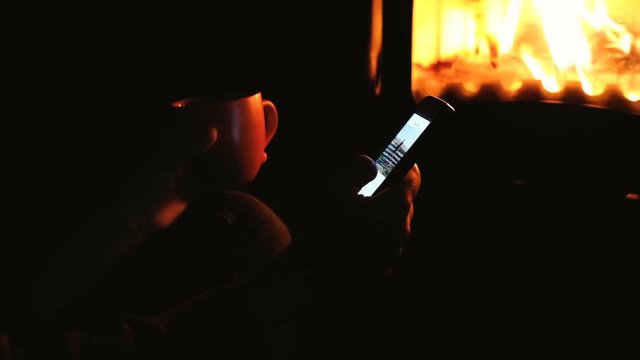 Girl resting by the fireplace and using smartphone for communicate in social networks. Close up of woman's hand holding cellphone and serfing internet in front of fireplace