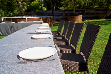 Fototapeta premium Dinner table with empty ceramic round plates and cutlery on grey tablecloth and chairs in garden outdoors, copy space. Dinner plate setting. White plate with knife and fork
