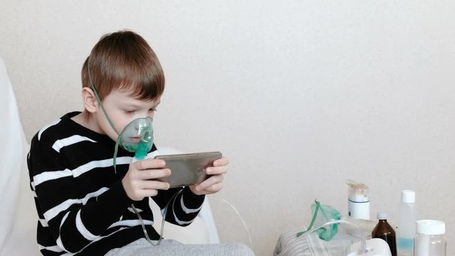 Use nebulizer and inhaler for the treatment. Boy inhaling through inhaler mask and playing the game in his mobile phone. Side view.