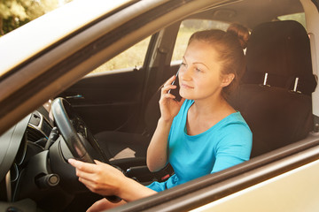 Beautiful young woman driving a car and talking on smart phone, free space. Business woman sitting in car with cell phone, looking on road, toned