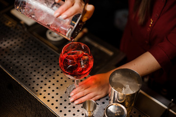 Bartender girl pouring a light red cocktail into the glass with ice
