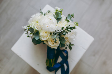 Close up A beautiful tender bridal bouquet in pastel colors of roses, peonies and green twigs tied with a long silk blue ribbon on background a wooden chair. Wedding accessories for bride