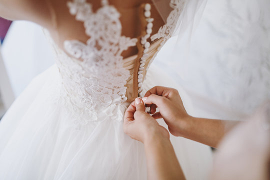 Close up Morning preparation newlyweds for the wedding. Female hands buttoning on a delicate lace pattern white dress with fluffy skirt on the waist of young slender bride. Wedding wear, accessories