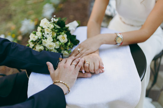 Beautiful wedding celebration. The groom in suit with bracelet and wedding ring and the bride in ivory dress with watch on her hand and bouquet of white roses holding hands on table with tablecloth