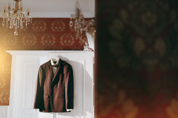 Beautiful wedding brown checkered jacket, a white shirt and a bowtie for the groom hang on a shoulders on the doorway to the room with beautiful patterned wallpaper. Wedding wear, accessories