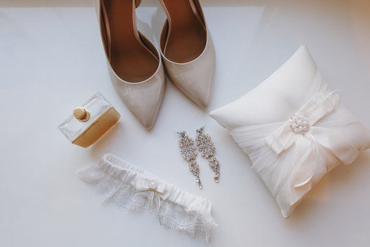 Close up Elegant light beige wedding varnish shoes with sharp noses, pillow for rings, earrings, perfume and garter for the bride on a white gloss windowsill background. Wedding wear, accessories